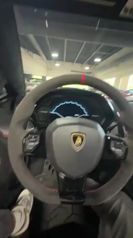 From the driver's seat, the video shows the interior of a Lamborghini, focusing on the steering wheel, dashboard, and center console. The steering wheel is gray with a red stripe at the top, and the Lamborghini logo is prominently displayed. The dashboard is black with red stitching, and the center console features a complex array of buttons and controls. The interior design includes carbon fiber elements. Outside the car, through the windows, other luxury cars can be seen, including a silver Porsche, indicating the scene is likely inside a dealership showroom.  The camera movement is slightly shaky as the person filming rotates the camera to showcase different parts of the car's interior. The roof of the car is partly open, letting in light and offering a clear view of the cars outside.