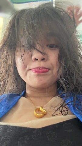 A close-up video shows a woman with wet, dark hair and bangs falling over her face. She has light skin, red lipstick, and a slight smile, with her tongue briefly out in the initial frames. She is wearing a gold necklace with two interlocking rings, a blue top or jacket, and what appears to be a light-colored top over a dark patterned one. Her hair appears tousled and messy, possibly being styled or dried. A hand briefly enters the frame to touch or adjust her hair. She closes her eyes and smiles contentedly as her hair is being worked on. The background is indistinct but appears to be an indoor setting with light-colored structures and some green accents. The location context provided is Quy Nhon Ward, Vietnam.