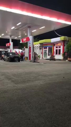 A nighttime shot of a Pertamina gas station with multiple cars and trucks queuing for fuel. A few employees in red shirts are visible, and the station lights illuminate the area. There are also a few motorcycles parked near the convenience store. The ground is paved, and the sky is dark.