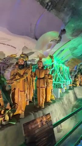The video shows the interior of a cave temple, brightly lit with colorful lights. Several statues of figures in orange robes stand in a row. A couple of people walk by. The video pans to show a large statue of a reclining Buddha, also illuminated with colorful lights. The cave ceiling is textured and painted in various colors, including green, white, and blue. A metal railing separates the walkway from the statue display.