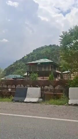 The video shows a passing view from a car window of homes and landscapes in a hilly area. The sky is cloudy, and the hills are covered in green trees and vegetation. The architecture of the homes varies, but generally includes fences and stone walls. There are also utility poles visible. The camera appears to be moving, following the road. Music is audible in the background.