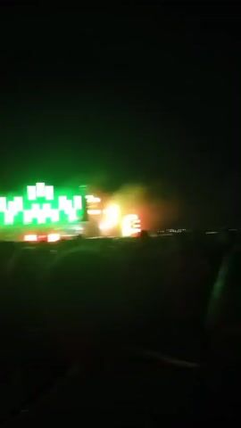 The video shows a crowd of people at an outdoor event at night. The stage is illuminated with bright lights, including a large screen displaying green geometric shapes. There is smoke or fog on stage, and bright lights are shooting upwards. In one shot, a tall, metallic structure with a red light at the top is visible, also illuminated by green lights. The sky transitions from dark blue to a gradient of orange and red near the horizon, indicating sunset or sunrise. People are seen from behind, silhouetted against the stage lights.