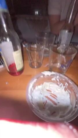 The video begins with a low-angle shot of a wooden table on which there are remnants of a gathering: bottles, glasses, an open container of a creamy salad, a pack of cigarettes, and other assorted items. The camera then pans to a poorly lit outdoor area where a young man in a black t-shirt and jeans sits in a chair, looking down with a serious expression. The lighting is dim, with the man's face partially illuminated.