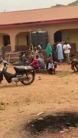 The video depicts a group of people gathered outside a yellow building with a red roof. Most of the people are seated on the ground in a shaded area, while some are walking around. Several children are also present, including one running. A motorcycle is parked in the foreground. The ground is dirt. Some of the women are wearing hijabs.