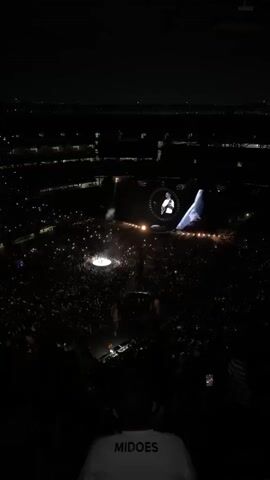 The video shows an aerial view of a concert at night. A large crowd of people is gathered in a stadium, illuminated by the lights from their phones. On the stage, a performer is visible on a large screen. The overall atmosphere is dark, with the focus on the stage and the crowd.
