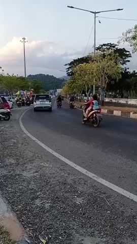 A paved road curves from the left side of the frame to the right. A gravel shoulder runs along the left side of the road, with a concrete curb bordering the road. On the left side of the road, several motorcycles are parked, and a white car is visible. Further in the distance on the left, people are standing near parked vehicles. On the right side of the road, a grassy area is separated from the road by a concrete divider with ornamental bushes. Trees with light green foliage line the right side of the road. A street light with multiple lamps stands near the center of the road on the right side. Several motorcycles are traveling on the road. One motorcycle, carrying two people, is in the foreground, moving away from the camera to the right. The sky is overcast with a soft, diffused light, suggesting it is late afternoon or early evening. In the background, a hilly, tree-covered landscape is visible.
