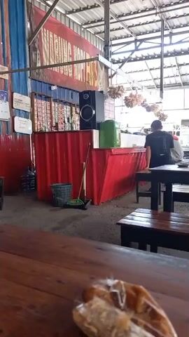 The video shows a dimly lit, open-air food stall or eatery. In the foreground, a blurry image of food, possibly bread, wrapped in plastic is visible on a wooden table. The background features a bright red counter constructed from a shipping container. Behind the counter, a black speaker sits atop a shelf displaying rows of packaged goods, likely snacks or cigarettes. To the left of the counter, a green garbage bin and a broom are present. Further back, two individuals are seated at a wooden table, their faces obscured. The environment appears to be covered by a corrugated metal roof, and the floor is concrete. The overall impression is of a casual, possibly roadside, establishment.