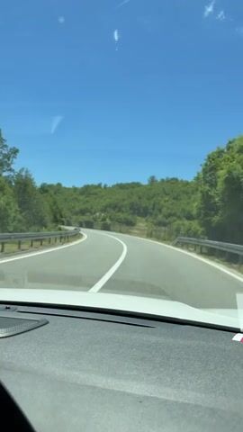The video shows a first-person view from a car driving on a paved road. The road is curving to the left, with white lane markings. The car is traveling through a hilly, green landscape with trees and some rocks. The sky is clear and blue with a few wispy clouds. The dashboard of the car is visible in the foreground. There are no people visible in the video.