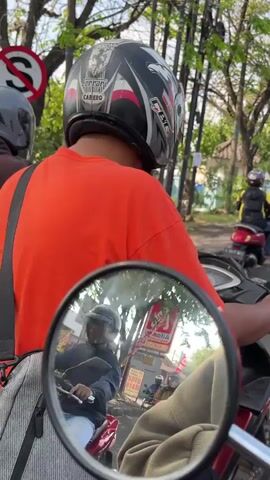 The video shows a person in an orange shirt and a helmet riding a motorcycle. They are in traffic, with other motorcycles and people visible in the background, some of whom are also wearing helmets. There are trees and a street sign visible. The scene appears to be outdoors during the day.