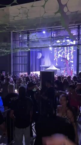 This is a vertical, low-angle video of a crowded indoor event. The lighting is dynamic with various colors and patterns. A DJ booth is visible in the distance with a DJ on a raised platform, illuminated by colorful lights. Several speakers are placed throughout the venue. People are standing and dancing in the crowd. Some individuals are facing the camera while others are looking towards the DJ. The ceiling is decorated with a network of green and white circular shapes. The overall atmosphere appears to be that of a nightclub or music festival.