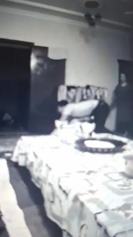 A blurry black and white video shows a room with furniture, including a bed or table covered with a patterned tablecloth. There are objects on the surface, like a plate, bowl, and cup. In the background, a woman in a dark dress is visible, standing near a clothes rack. The video is shaky, and at one point, the camera is moved abruptly, resulting in a bright, overexposed image of a shoulder, possibly with a red mark or stain.