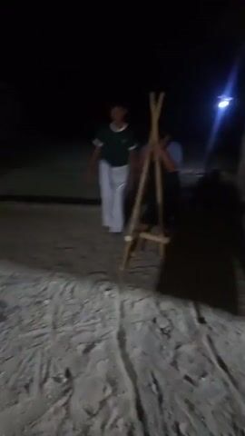 The video shows two young boys in Kendel, Indonesia, at night, possibly playing. The boy on the left is wearing a dark green t-shirt and light gray pants, standing on the sandy ground. The boy on the right is wearing a blue and black t-shirt and dark shorts, and is standing on stilts made of bamboo poles. He is holding onto the stilts with both hands and appears to be in the process of walking or balancing. The lighting is dim, suggesting it's nighttime, and the ground appears to be a mixture of sand and dirt, typical of a beach or rural area. The overall mood seems playful and adventurous as the boy on stilts navigates his environment. No visible text is present in the video.