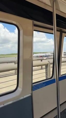 The video shows a view from inside a train at an airport. The train is moving, and through the window, one can see the airport runway and several airplanes parked on the tarmac. The sky is blue with scattered clouds. The train has a white and grey interior with metal handrails. There is a sign on the door in multiple languages, including English, that reads, "Please do not lean on doors." A male voice can be heard making an announcement, likely the train operator.
