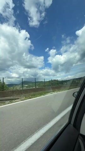 The video is taken from inside a car, looking out the passenger window. The car is driving on a paved road with a white line on the right side. To the left of the road, there is a grassy area, followed by a green fence and then rolling hills covered in vegetation. The sky is blue with scattered white clouds and a faint contrail. A white Subaru Forester passes the car. Further down the road, there is a body of water with some buildings on the far shore. The interior of the car is visible in the foreground on the right side of the frame, showing part of the door and window frame.