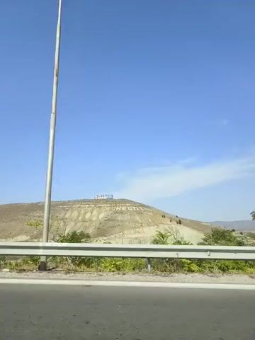The video shows a view from a moving vehicle, looking towards a large hill covered in vegetation and some signs at the top. The sky is clear and blue. The vehicle is traveling on a road with a guardrail in the foreground. There are some trees and bushes along the side of the road. The signs on the hill appear to have text on them, but it is difficult to read clearly. The overall impression is of a sunny day in a semi-arid landscape.