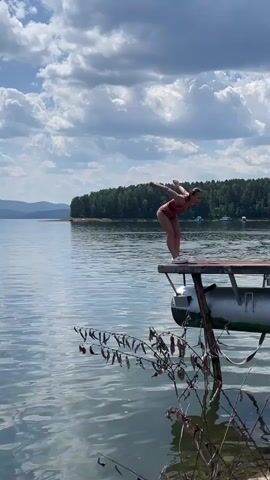 A person in a red swimsuit jumps backward from a wooden dock into a large body of water. The person's body is arched in mid-air. The dock has metal pontoons underneath it. The water is calm and reflects the blue sky and white clouds. A dense green forest and distant mountains are visible on the opposite shore. There is vegetation in the foreground.