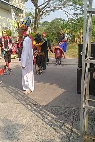A group of people in colorful traditional costumes are walking in a procession on an asphalt surface outdoors. They are carrying spears and wearing elaborate headpieces and masks. Some people in the background appear to be watching the procession. There are trees and buildings visible in the background, suggesting an outdoor event or festival. The sun is casting shadows on the ground.