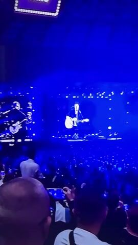 The video shows a concert with a performer on stage playing a guitar and singing. The stage is lit with bright blue lights, and there are many small white lights in the background that appear to be part of the stage design or lighting effects. A large crowd of people are visible in the foreground, many of whom are holding up their phones to record the performance. The audience is densely packed, and the overall atmosphere appears to be energetic and engaging. The performer is wearing dark clothing and a hat, and is standing on a raised platform. The screen behind the performer is displaying a silhouette of the performer, creating a striking visual effect.