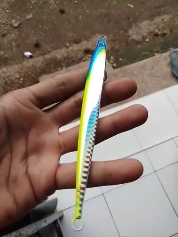 Here is a brief description of the video:

The video shows a person's hand holding a slim, metal fishing lure. The lure is brightly colored with blue, yellow, green, and rainbow stripes, with a checkered pattern along its length. It has loops at both ends for attaching to fishing lines. The background includes tiled flooring and a glimpse of an outdoor area.