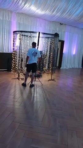 A wide shot pans across a large, empty event space with a wooden floor. The ceiling is draped with white fabric, illuminated by colored lights that shift between purple and blue. The walls are also lined with white fabric and accented with vertical colored lights. On the left side of the room, a man in a white t-shirt and black shorts stands with his back to the camera, facing a backdrop of string lights. Further into the room, several round tables draped in white cloths are set up, with chairs around them. In the background, near a large window or open area, a few people are visible, including one person in a white dress. The overall impression is a venue prepared for an event, with decorative lighting and seating arrangements in place.