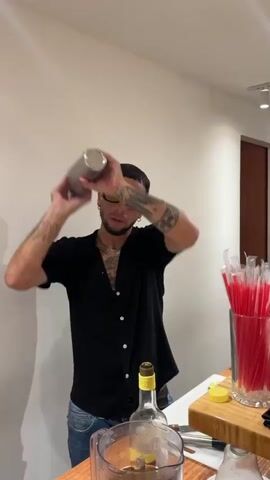 A person with yellow-tinted glasses and tattoos is shaking a metal cocktail shaker. He is wearing a black button-down shirt and jeans. Behind him, on a white surface, are various bottles, red straws in a glass, and sliced limes. The background is a plain white wall.