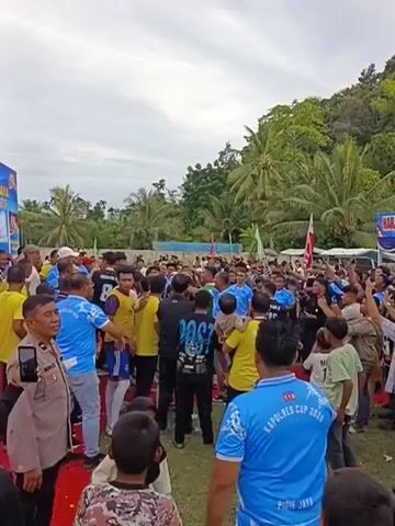 A large group of people are gathered outdoors, possibly at a sporting event. Many are wearing blue t-shirts with "KAPOLRES CUP 2020" visible on the back of one. Others are wearing yellow t-shirts. There is a banner in the background with Indonesian text and images of trophies, suggesting an award ceremony or tournament. A police officer in uniform is visible on the left side of the frame. Some individuals are holding up their hands or cheering, indicating excitement or celebration. The environment appears tropical, with palm trees and lush greenery in the background. The sky is overcast.