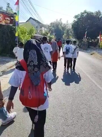 The video shows a group of people walking down a paved road in a rural or suburban area. The sun is shining, casting long shadows. The people are diverse in age, with many appearing to be teenagers or young adults. Some are wearing white t-shirts with graphics, while others are wearing red shirts or casual clothing. One person in the foreground is wearing a black hijab and a red backpack. There are houses and trees visible on either side of the road, and utility poles with wires run along the side. A motorcycle is seen in the background. The atmosphere seems to be that of a procession or a group outing.