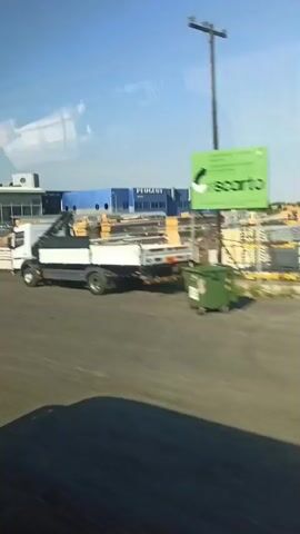 The video shows a view from inside a moving vehicle, looking out at an industrial or commercial area. On the right, a white truck with a crane is visible, parked or driving. In the background, several buildings are present. One prominent building is a large, bright blue structure with the word "PEUGEOT" in white lettering on its facade. Adjacent to it is another building, white with a modern design, featuring large glass windows. In front of these buildings, there are stacks of what appear to be building materials or pallets, some with yellow wrapping. The area in the foreground is a gray, paved surface, possibly a road or parking lot, with the shadow of the vehicle the camera is in cast upon it. The sky is clear and blue. As the video progresses, the view shifts slightly, revealing more of the buildings. Some appear to be car dealerships with brand names like "ISUZU" and "SUBARU" visible. The overall impression is of a business park or industrial estate.