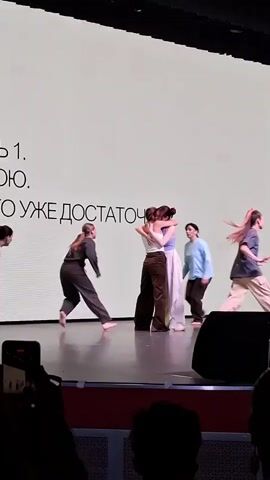 On a stage, a group of dancers are performing. In the center, two women embrace. They are surrounded by other dancers moving around them. The stage is brightly lit, and behind the dancers is a large screen with text on it in a language other than English. In the foreground, the heads of audience members are visible, along with some phones recording the performance.