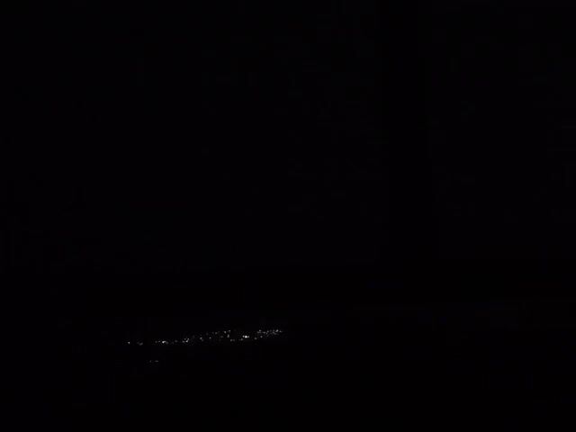 The video is shot at night. The scene is mostly dark, but in the distance is a faint array of lights suggesting a town or city. The lights are spread out in a line. There are no people, objects, actions, or interactions visible.