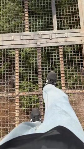 The video begins with a close-up shot of a person's legs and feet standing on a metal grate walkway. The camera then pans to the right, revealing a long, narrow walkway with metal railings and a grated floor, overlooking a lush green forest and valley below. The scene then shifts to a wider shot of the surrounding mountainous terrain, with trees, rocks, and a visible structure on a distant hillside. The video captures the perspective of someone walking on an elevated walkway, offering panoramic views of a natural landscape.