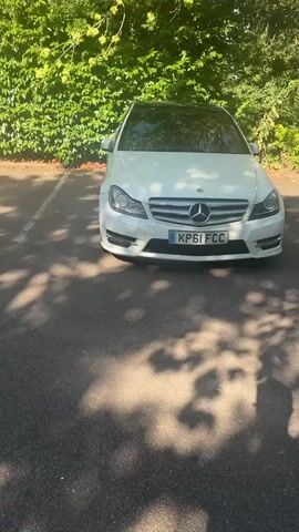 A white Mercedes-Benz C-Class W204 is parked on asphalt. The car is facing forward, with its license plate visible. Sunlight filters through tree leaves, casting shadows on the ground. A green, leafy hedge is behind the car. The video shows a slight, slow pan to the right.