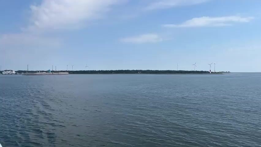 The video shows a wide view of the ocean with a distant shoreline. On the shoreline, there are several wind turbines, some buildings, and what appears to be a harbor or industrial area. The water is calm with small ripples, and the sky is blue with scattered white clouds. The view seems to be from a boat or ship, as indicated by a white railing in the foreground on the left side of the screen. The camera pans slowly to the right, revealing more of the water and the distant horizon. The overall scene is peaceful and suggests a sunny day at sea.
