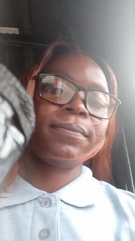The video begins with a close-up of a Black woman wearing glasses and a grey collared shirt. She is looking around, and her hair is a reddish-brown color. The camera then pans to the right, showing a street scene with buildings, trees, and a cloudy sky. A pink and white poster with images of people is visible on the left side of the frame. Further down the street, a man in a blue shirt is standing near a power pole. The video ends with a view of houses and a fence along the side of the road.