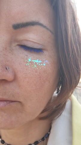The video shows a close-up of a person's face.  The person is facing forward, but oriented slightly towards the left.  The person's left eye is open, and it is ringed with blue mascara.  There is also green and light blue glitter on the person's left cheek, below the eye.  The person has a pierced nose, and freckles on the face. The person also wears a beaded necklace.  The person has brown hair.  The person appears to be outside in daylight.