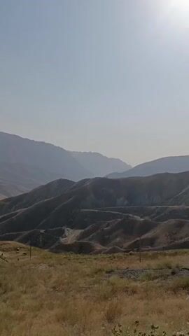 This is a vertical, handheld video that pans across a dry, mountainous landscape under a clear sky. In the foreground, there is dry grass and a patch of gravel. As the camera pans, it reveals rolling hills with sparse vegetation and a few scattered trees. In the distance, larger, more rugged mountains are visible. The overall impression is one of a vast, arid, and sparsely populated region. There are no people or discernible actions taking place in the video. The lighting suggests it is daytime, likely late morning or early afternoon, with the sun casting shadows that accentuate the contours of the hills. There is some litter visible in the foreground, suggesting human presence, but it is not the focus of the video.