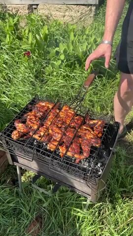 Here's a description of the video:

A person wearing black shorts and a bracelet on their wrist is grilling meat on a small, portable charcoal grill. The grill is rectangular and made of black metal. It is placed in a grassy area next to a building with a wooden base and white wall. The person lifts the grilling rack, which is full of what appears to be chicken pieces marinated in a red sauce, to show the lit charcoal underneath. They then lower the rack back onto the grill. The grill is surrounded by tall green grass.