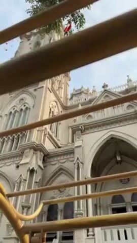 The video shows the view from inside a yellow metal structure, possibly a bus, looking out at a large, ornate, light-colored church with a tall tower.  The camera pans up, revealing the full height of the tower, decorated with architectural details like arches and windows. A few sparse tree branches and strings of colorful pennants are visible at the top of the frame against a blue sky. The camera is slightly shaky, suggesting it is being held by someone in motion. The overall environment seems calm and sunny. The church's architectural style appears to be Gothic or neo-Gothic.