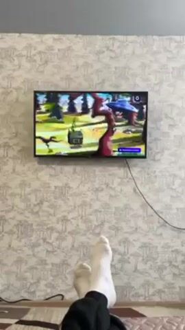Here's a description of the video:

A person is lying on a bed with their feet up, watching television. The TV screen shows a cartoon. The walls have wallpaper with a subtle tree pattern. A rug with dark brown and maroon blocks is seen on the lower left. The person is wearing white socks and black pants.