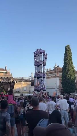 A human tower is being built in a public square. The tower is composed of many people stacked on top of each other, forming multiple levels. The people in the tower are wearing matching purple shirts with white trim and white pants. The tower is quite tall, reaching several stories high. A large crowd of spectators surrounds the base of the tower, many looking up at the impressive display. Some people in the crowd are holding cameras or phones, presumably to capture the event. In the background, there are buildings, trees, and a clear blue sky. The scene appears to be taking place outdoors during the daytime.