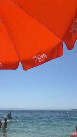 The video shows a vibrant orange umbrella with white text, "APEROL SPIRIT", partially obscuring a clear blue sky and the ocean. The umbrella is positioned in the foreground, angled downwards towards the viewer. Below the umbrella, people are visible in the turquoise water. Some are swimming or wading, while others appear to be standing or playing. In the distance, the ocean stretches to the horizon, meeting a hazy landmass. The overall scene evokes a sense of a sunny day at a beach or seaside resort.