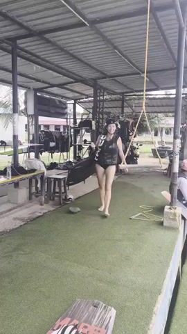 A woman wearing a black life vest and helmet, and a black swimsuit walks barefoot across a green artificial turf mat. She is carrying a black wakeboard. She turns and smiles towards the camera as she walks. Behind her, a body of water is visible with a wakeboarding cable system overhead. Other people, wearing life vests and helmets, are also present near the water and are preparing to wakeboard. The environment appears to be an outdoor recreational facility.