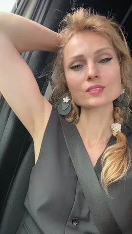 A blonde woman with her hair in braids and wearing a black vest and seatbelt is in the passenger seat of a vehicle. She is looking at the camera and touching her hair. The window shows a green field with trees in the background.
