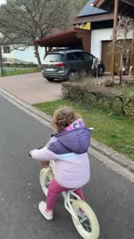 A young child with curly hair rides a white balance bike on an asphalt street. The child is wearing a pink and purple jacket with a light blue stripe, pink pants, and white sneakers. They ride towards the camera and look to the side with a smile. A woman in a black Nike sweatsuit and Converse sneakers walks behind the child. A gray SUV is parked in a driveway near a modern house with solar panels on the roof. The setting appears to be a residential area with trees and greenery visible.