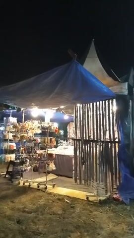 The video shows an outdoor scene at night, likely a market or fair. A large tent with a blue tarp roof is visible, with stalls set up underneath. One stall appears to be selling accessories, with various items displayed on racks. Another stall is adorned with a large banner advertising "Sweet Ice Cream" and features images of ice cream cones. A person wearing a hijab walks towards the camera in front of the ice cream stall. The ground is covered with dry grass and straw. The lighting is dim, with artificial lights illuminating the stalls.