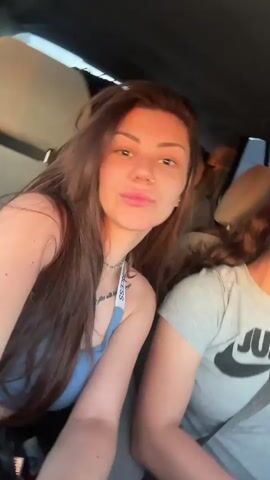 The video shows two women in a car. The woman on the left has long brown hair and is wearing a light blue top. The woman on the right has her hair pulled back and is wearing a grey t-shirt that says "Just Do It" with a Nike logo. The woman on the left leans in and kisses the woman on the right on the cheek. The woman on the left then smiles at the camera. The background shows the inside of a car with luggage in the back. The scene appears to be filmed from the passenger seat.