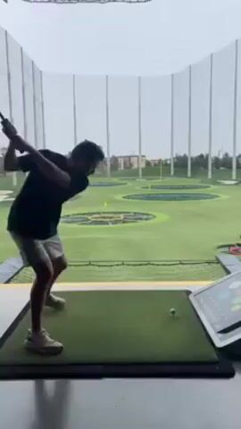 A man is seen hitting a golf ball at a Topgolf location. The man is facing away from the camera, wearing a black t-shirt, light gray shorts, and white sneakers. He swings the golf club and watches the ball fly towards the green targets in the distance. The Topgolf venue is visible in the background, complete with netting, artificial turf, and a digital screen showing game information. The weather appears overcast.