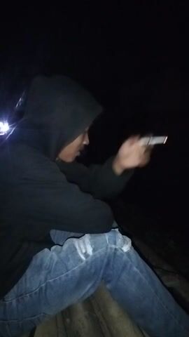 The video begins with a close-up of a person in a hooded jacket and jeans, holding a cigarette. The scene appears to be outdoors at night, with darkness dominating the frame. The camera then pans upwards to reveal a white truck with illuminated headlights and taillights, suggesting it is either parked or moving slowly. The view shifts again to show the interior of what appears to be a bus or van, with green and red lights illuminating the space. The final frames show brief, blurry glimpses of people and lights, making it difficult to discern specific details. The overall impression is of a dimly lit, potentially urban or roadside environment, with individuals engaged in casual activity, possibly related to transportation or a gathering.