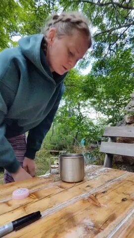 A woman with her hair in a braid, wearing a dark green hooded sweatshirt and a striped skirt, is pouring water from a large plastic bottle into a metal mug that sits on a wooden table. The table also has a pink object and a black marker on it. The background shows greenery and trees.