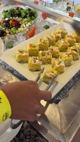 A person is using tongs to take a serving of yellow layered food from a tray on a buffet line, placing it onto a white plate. The food appears to be cut into small square or rectangular pieces, topped with white and green, possibly herbs. There is a salad to the left of the tray on a bed of ice. The person is wearing a yellow wristband. The buffet line looks like it is made of stainless steel with a marble-like counter.